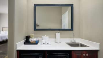A hotel bathroom with a white countertop, a sink, a mirror, a coffee maker, and a dish soap dispenser.