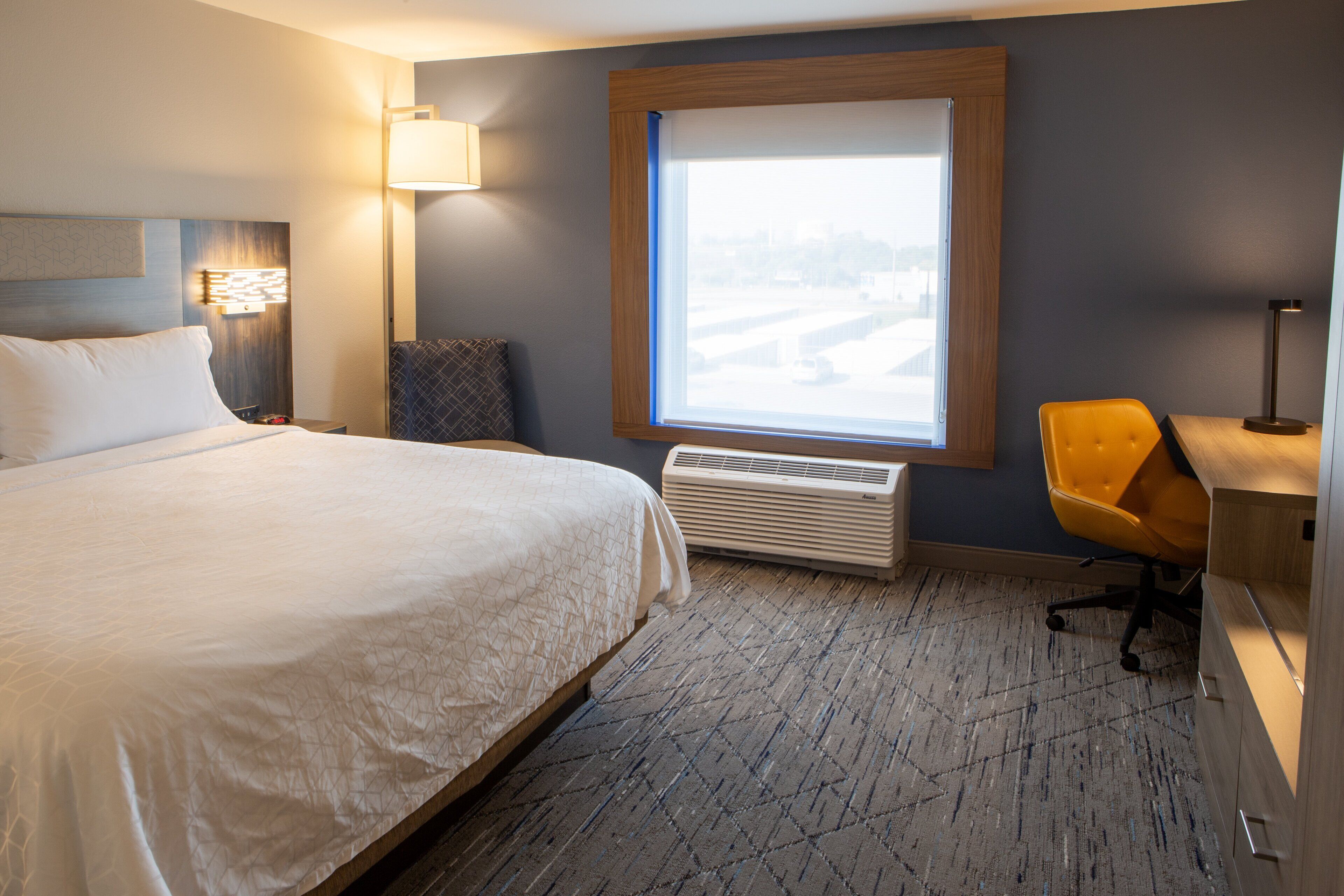 Una habitación de hotel con una cama grande, un escritorio con una silla, un ventanal con vistas y un aire acondicionado instalado en la pared.