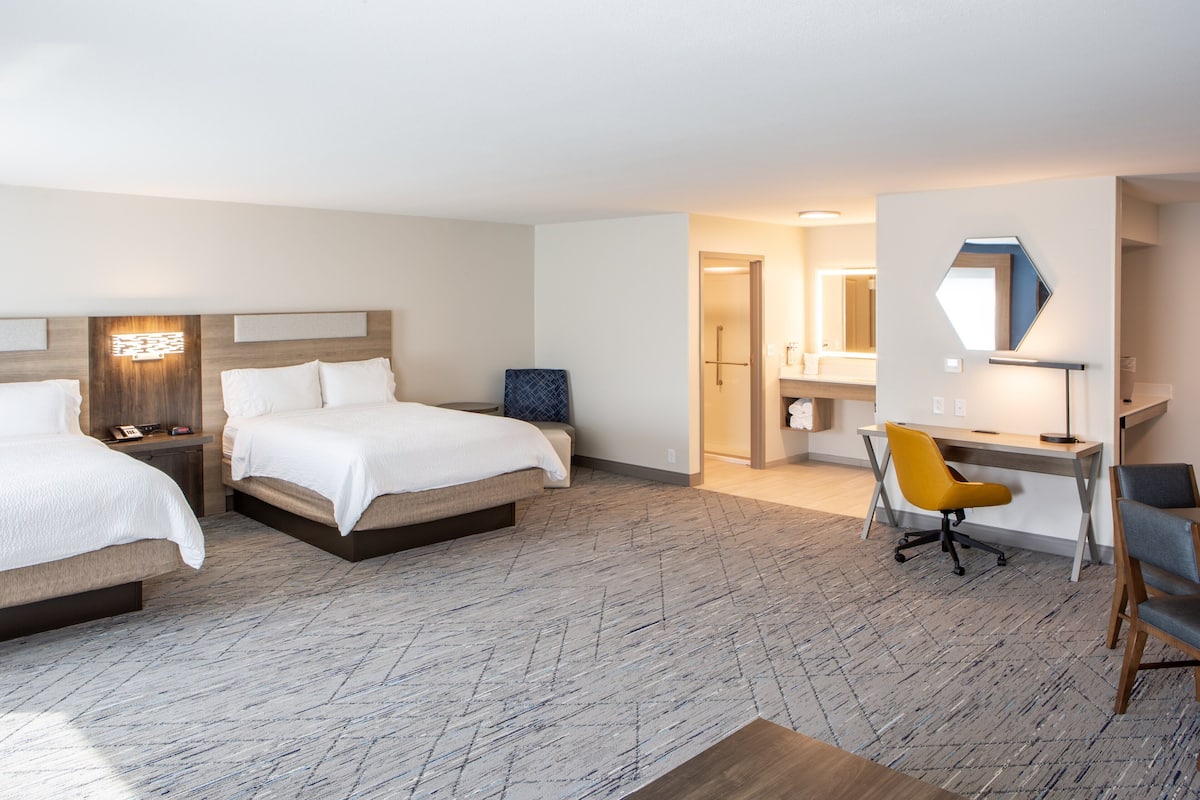 Une chambre d’hôtel avec deux lits, un bureau, une chaise et une salle de bain visible à travers une porte ouverte.