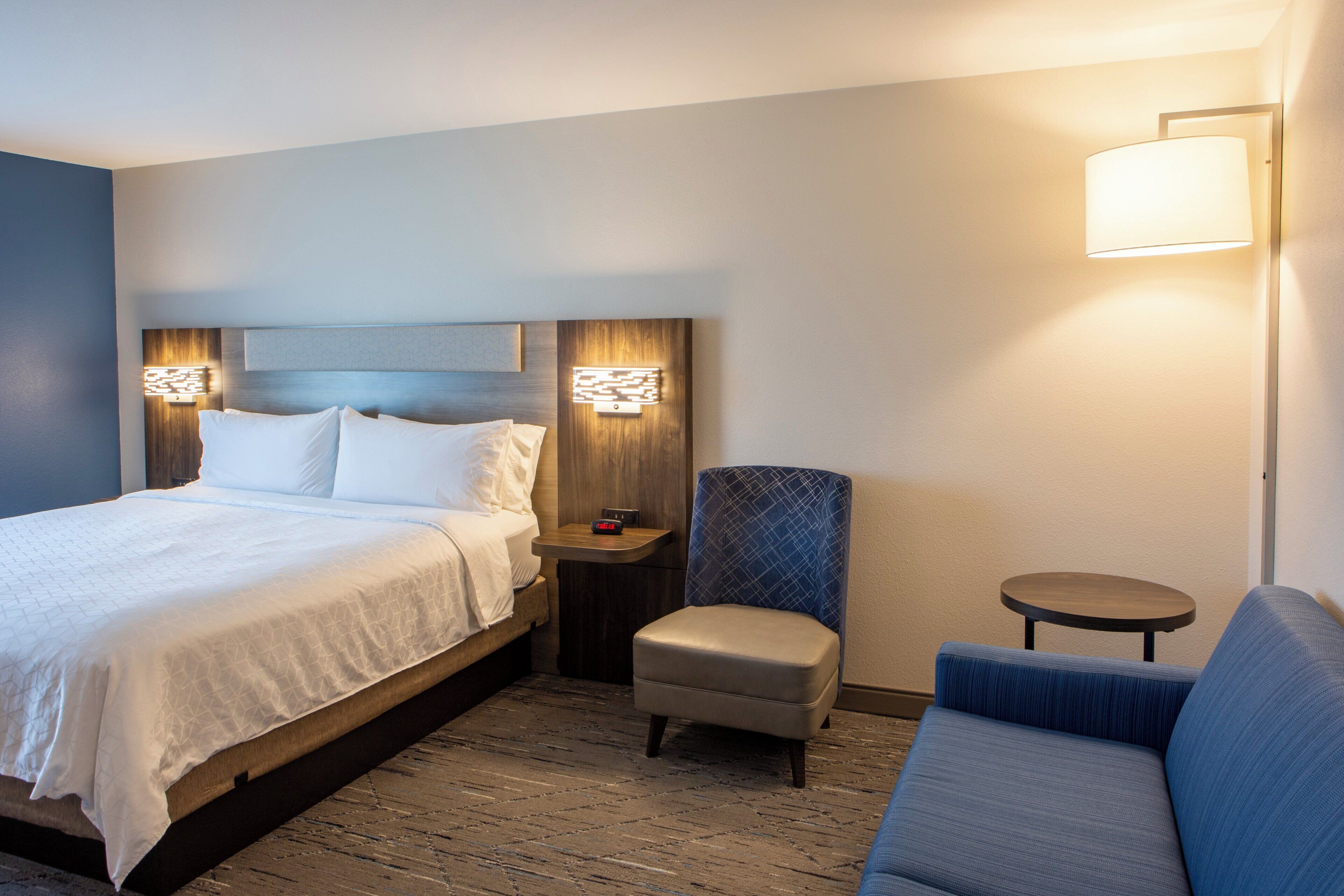 Une chambre d’hôtel avec un lit, une chaise, une petite table et une lampe fixée au mur.