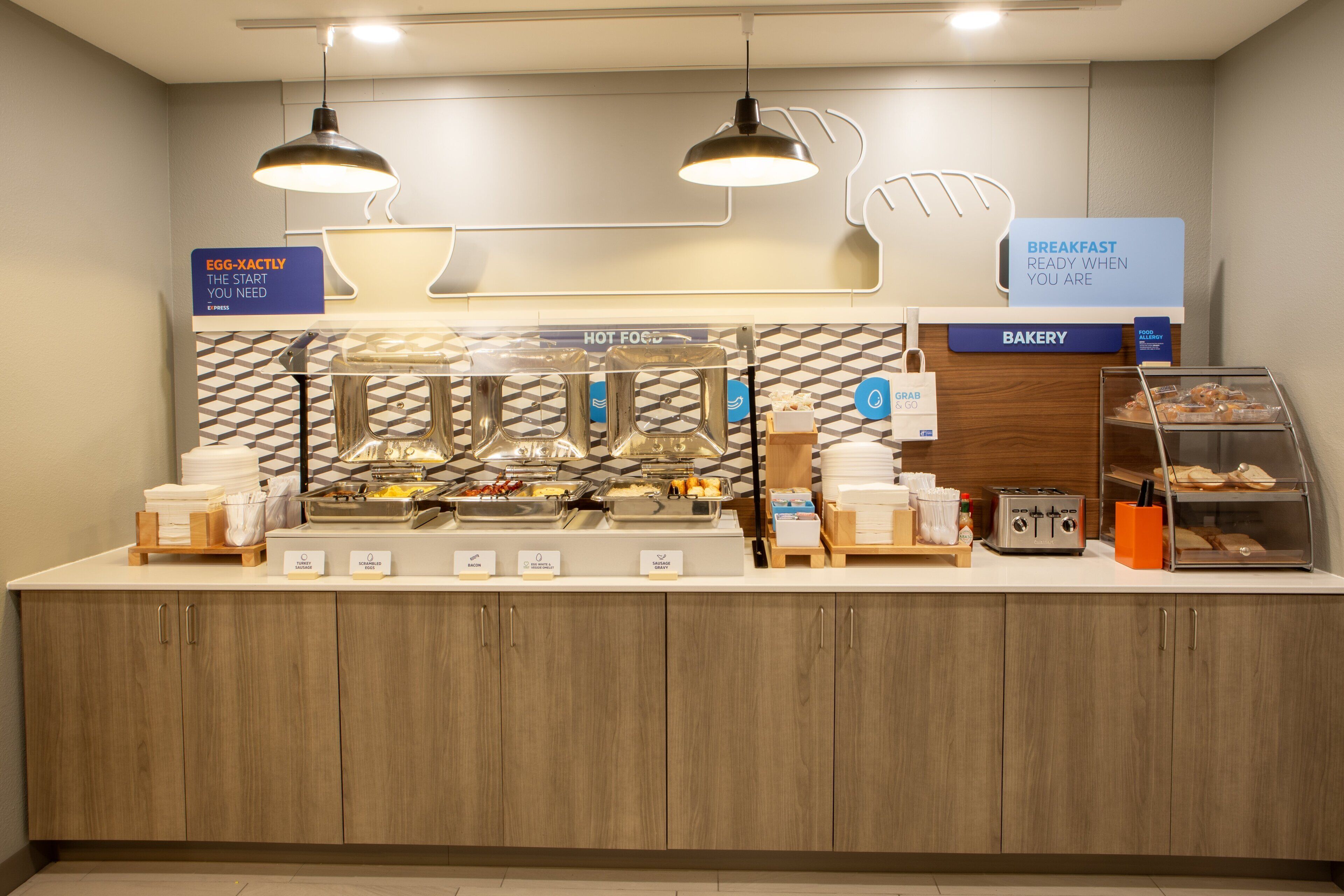 Zona de desayuno tipo buffet con estaciones de comida señalizadas, una sección de panadería y un letrero de comida para llevar.