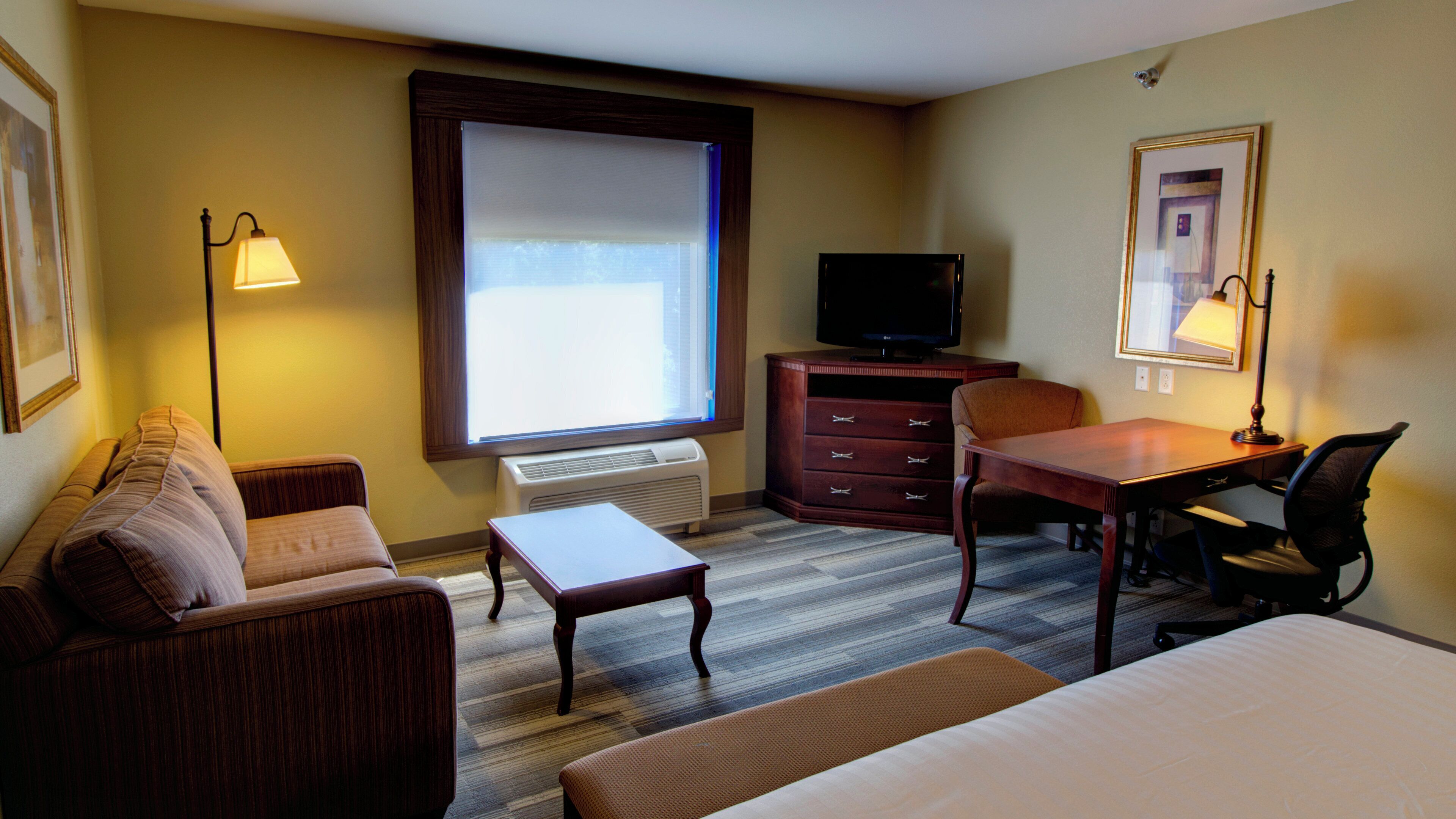 Quarto de hotel com cama, escrivaninha, poltrona, sofá, televisão e janela com persianas.