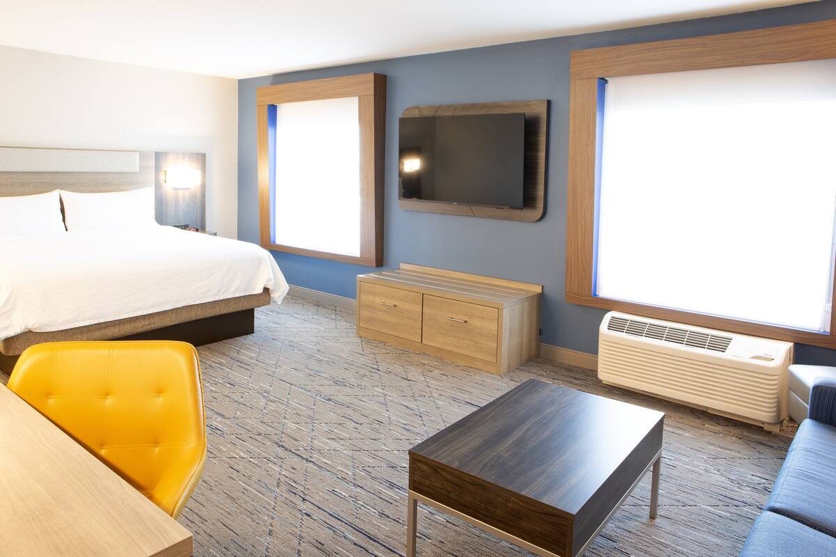 Une chambre d’hôtel équipée d’un lit, d’un bureau, d’une chaise, d’une commode, d’une télévision et d’une fenêtre avec des stores.