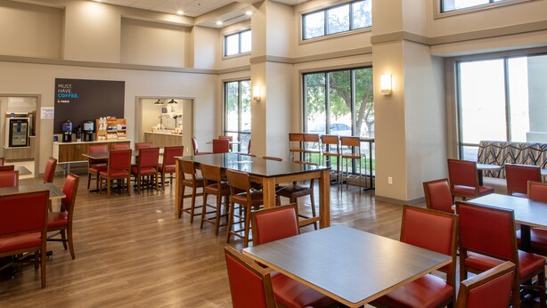 Un vaste espace dédié à la salle à manger, avec de nombreuses tables et des chaises rouges, de grandes fenêtres laissant entrer la lumière naturelle, et un coin café à l’arrière-plan.