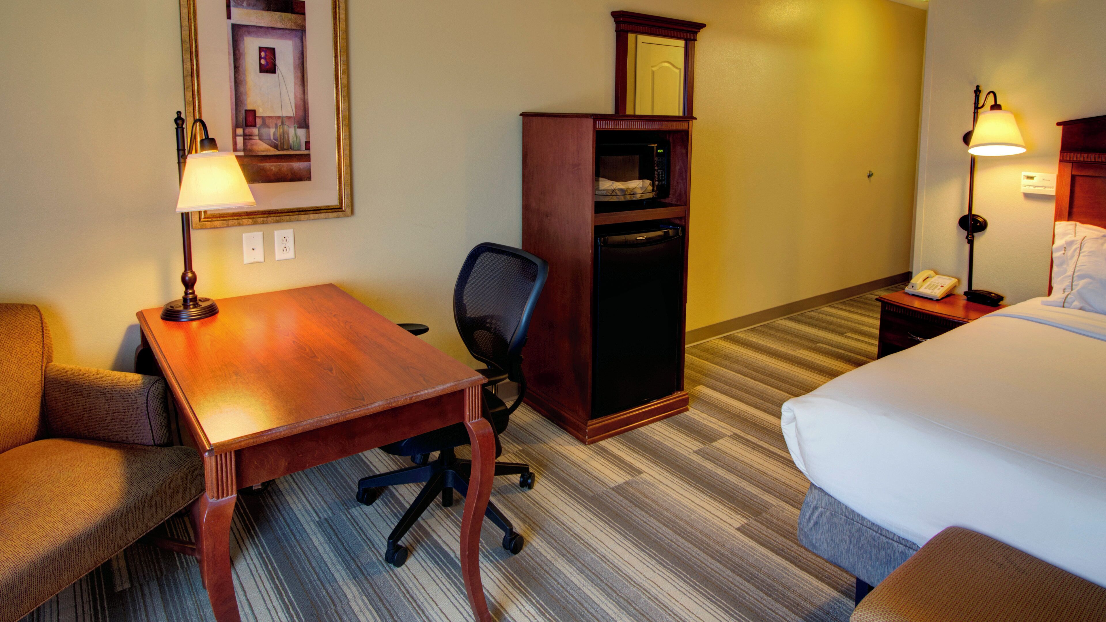 A hotel room with a wooden desk, a chair, a bed, a wardrobe, and a nightstand with a lamp.