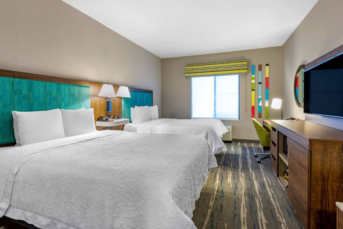 A hotel room with two beds, a desk, a chair, a TV, and a window with curtains.