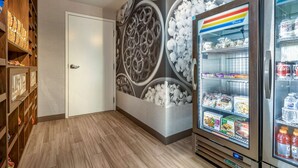 A hotel room with a wooden shelf unit, a door, and a wall with a circular pattern. There are two refrigerators with various food items and beverages. - Hampton Inn Carlsbad-North San Diego County (Carlsbad)