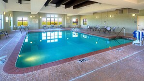 An indoor swimming pool with clear water, surrounded by lounge chairs and tables, and large windows offering a view of the outdoors.