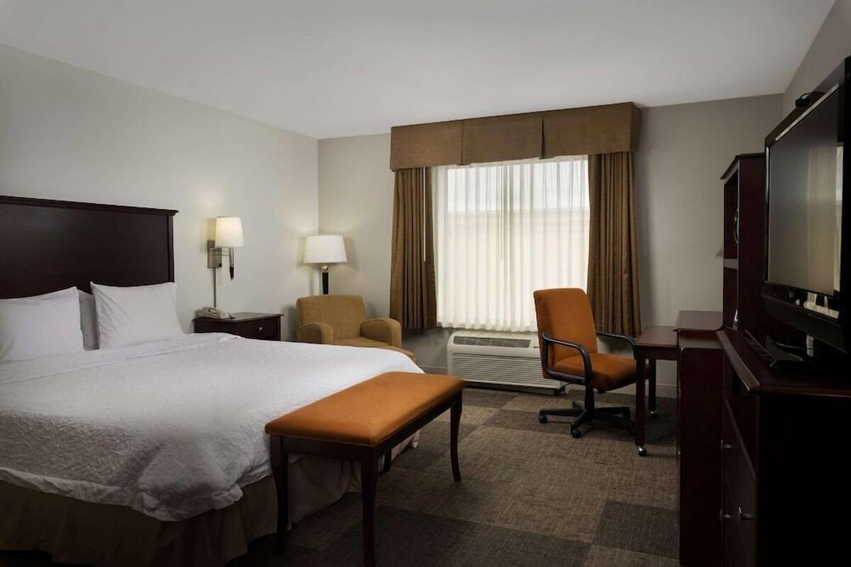 Habitación de hotel con una cama grande, un escritorio con televisor, una silla, una lámpara y un ventanal con cortinas.