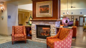 A lobby with a fireplace, two patterned armchairs, a large decorative vase, and a framed picture above the fireplace.