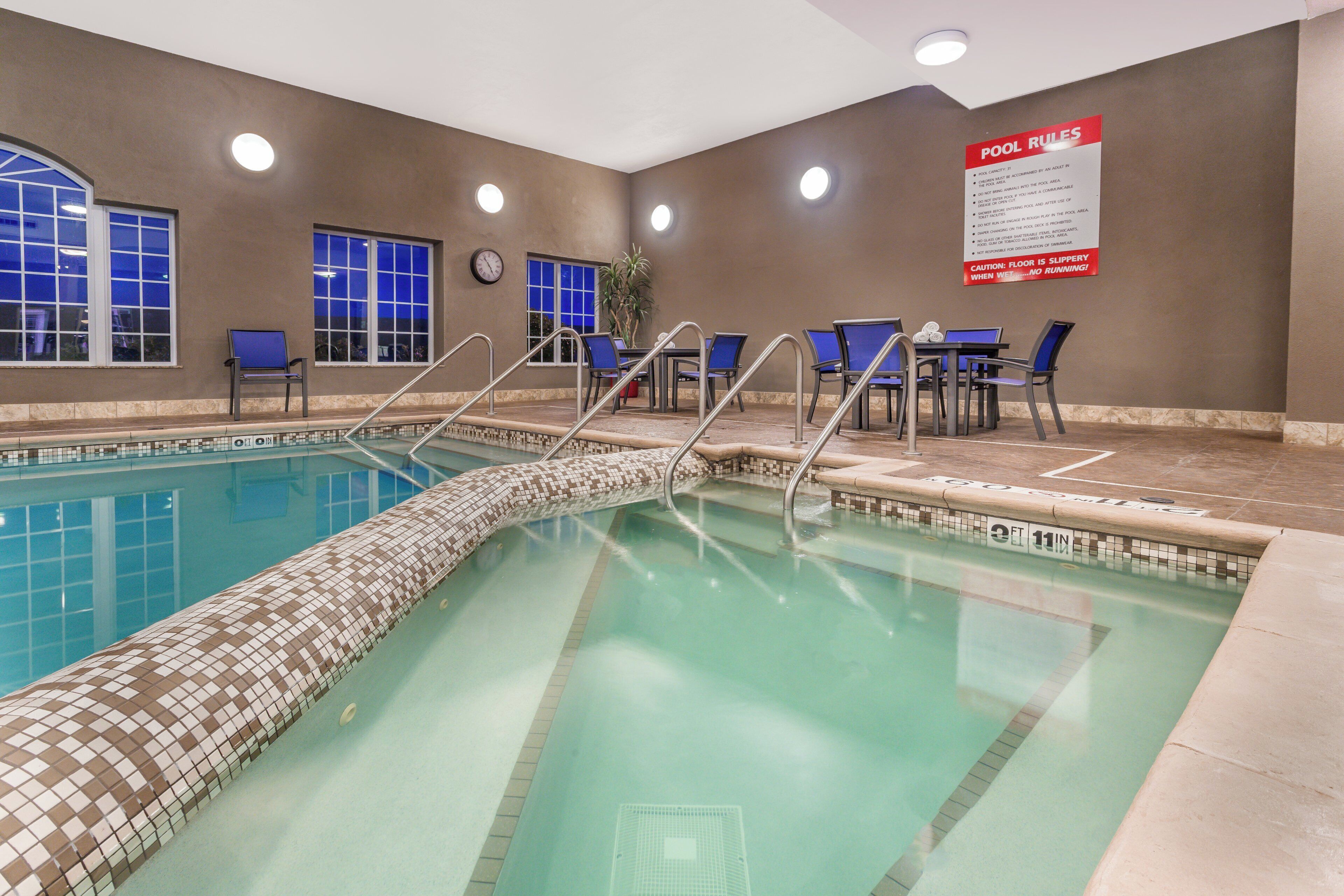 An indoor swimming pool with a tiled edge, a pool rule sign, and seating area with chairs and tables.