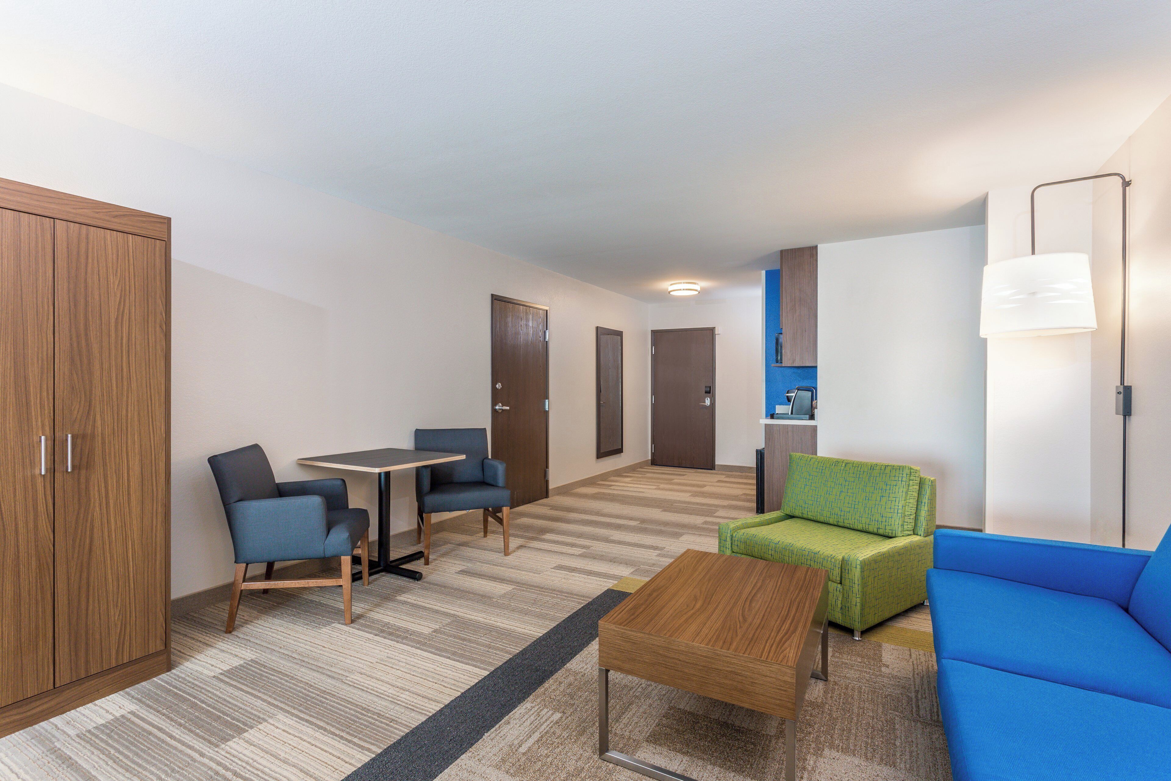 Una habitación de hotel con un armario de madera, una mesita con dos sillas, un sofá verde y un sofá azul, además de una lámpara.