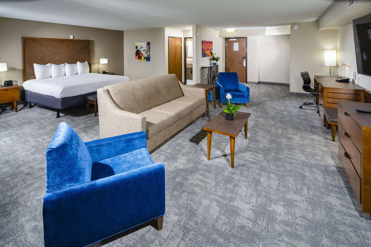 A hotel room with a large bed, a sofa, a blue armchair, a desk, and a television.