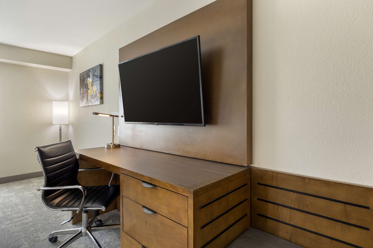 A hotel room with a wooden desk, a black office chair, a flat-screen TV mounted on the wall, and a painting on the wall.