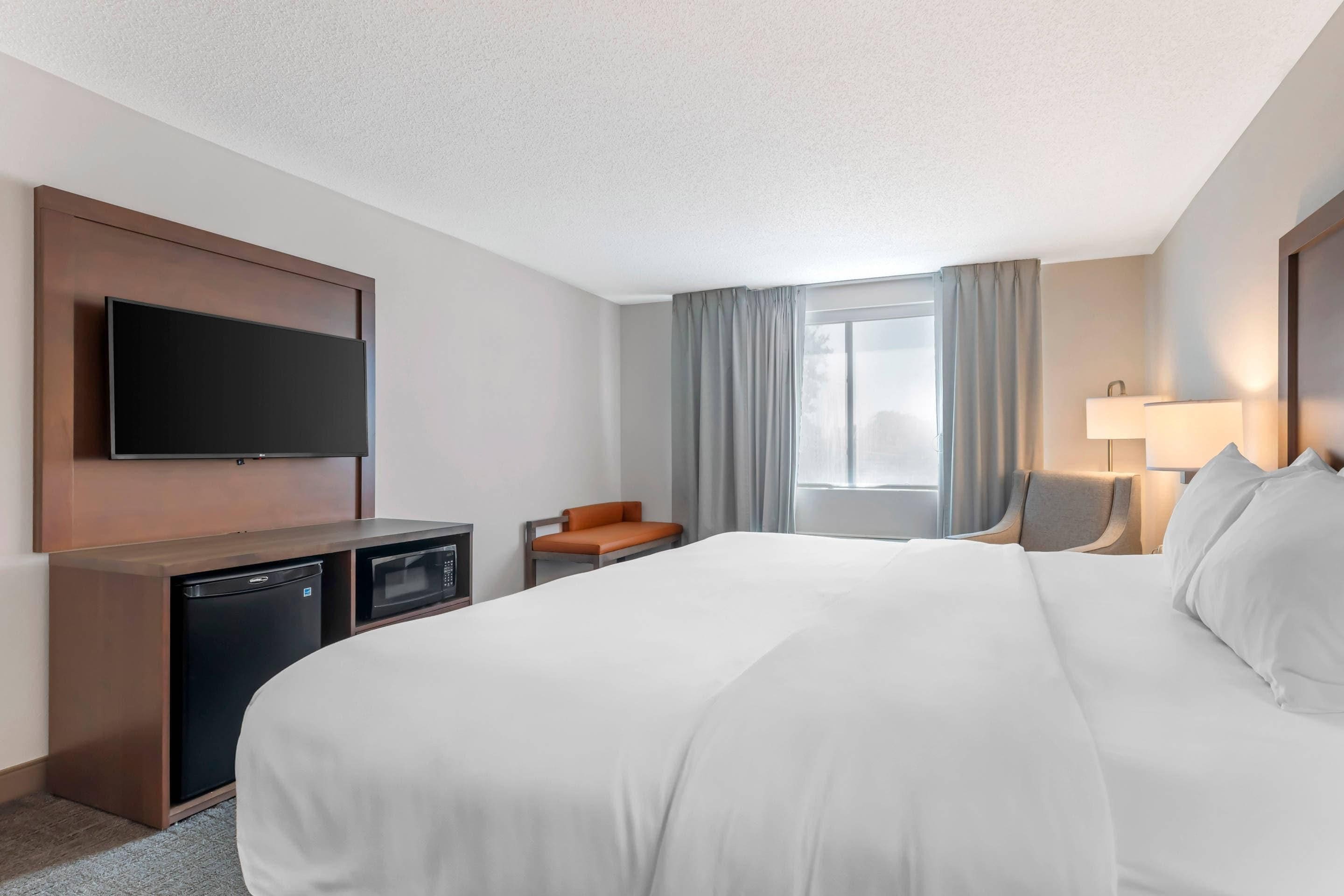 A hotel room with a large bed, a flat-screen TV mounted on the wall, a desk with a microwave, and a chair.