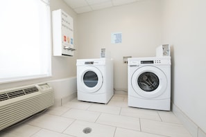 A laundry room with two front-loading washing machines, a wall-mounted air conditioner, and a sign indicating laundry products are available for purchase.