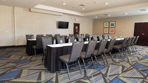 A conference room with rows of tables and chairs, a flat-screen TV mounted on the wall, and a door with a security camera.