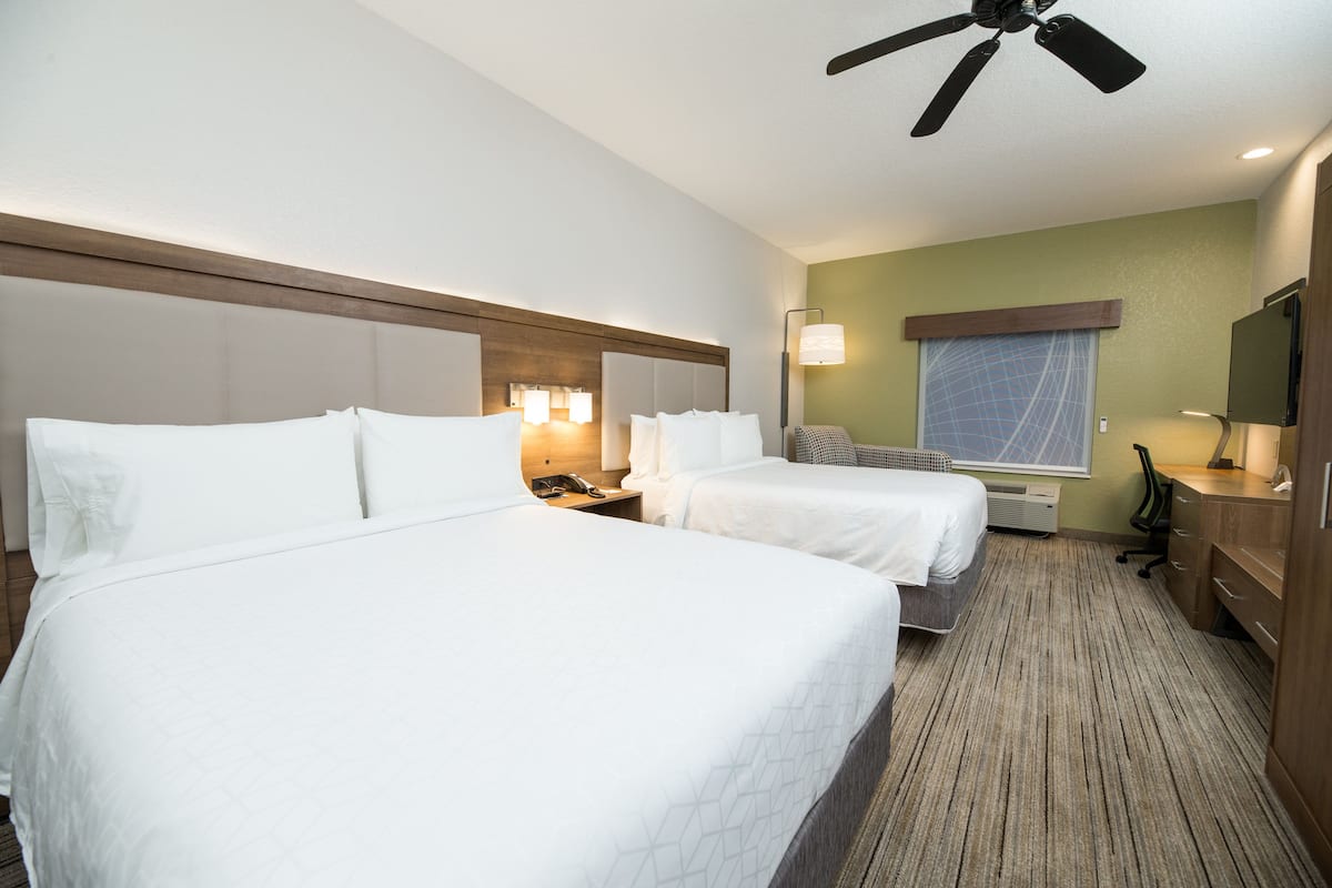 A hotel room with two beds, a ceiling fan, a TV, a desk with a lamp, and a window with a view.