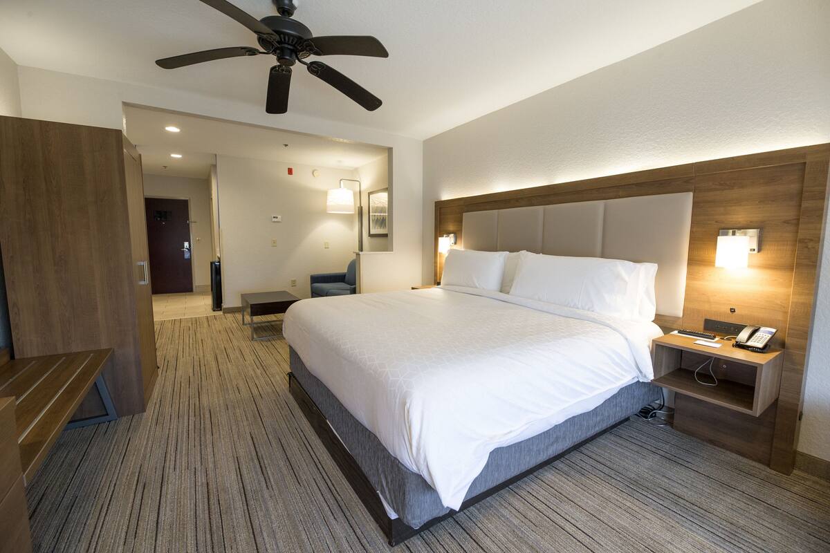 A hotel room with a large bed, a desk, a chair, a ceiling fan, and a nightstand with a phone.