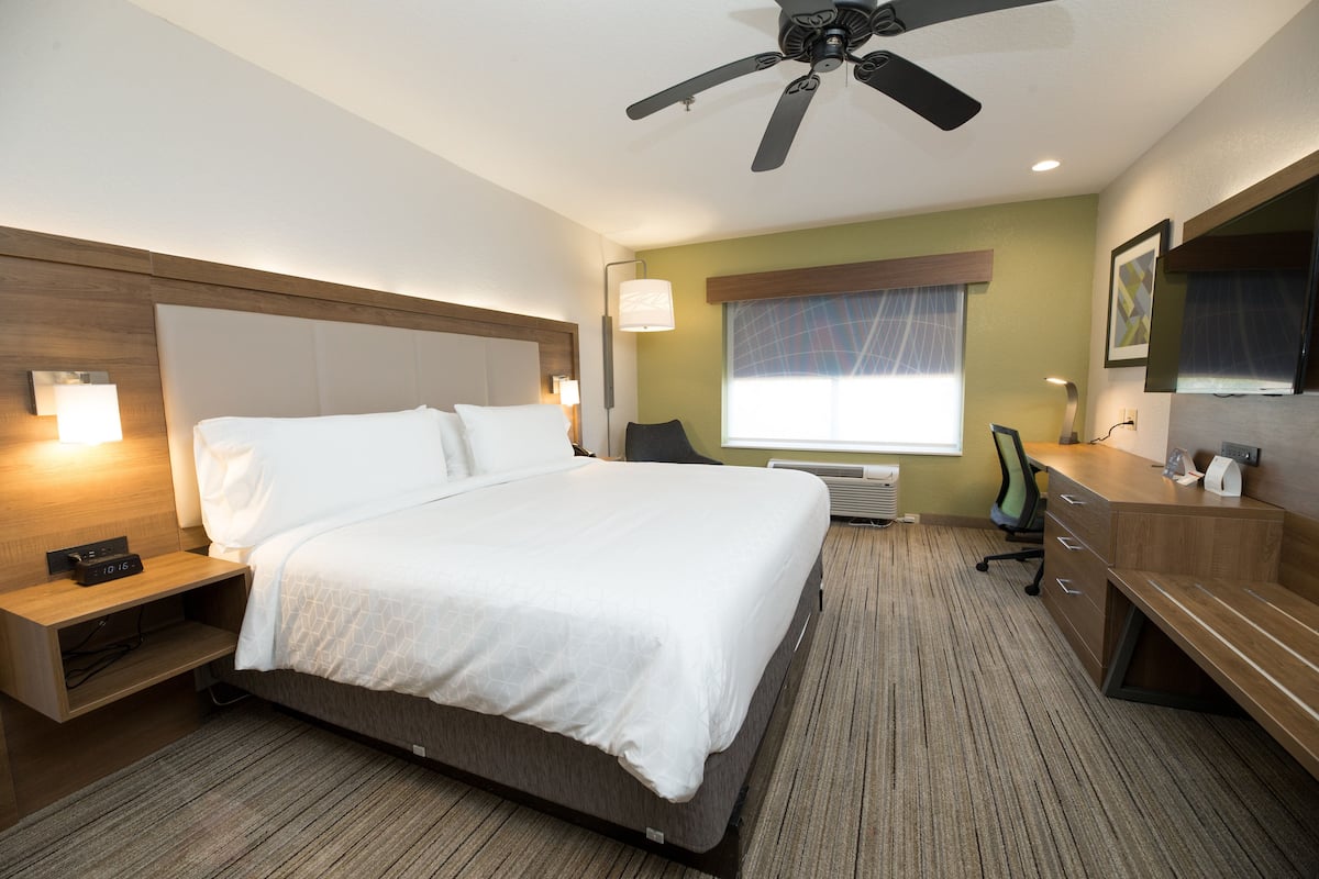 A hotel room with a large bed, bedside tables, a desk, a chair, a ceiling fan, a window with curtains, and a wall-mounted TV.