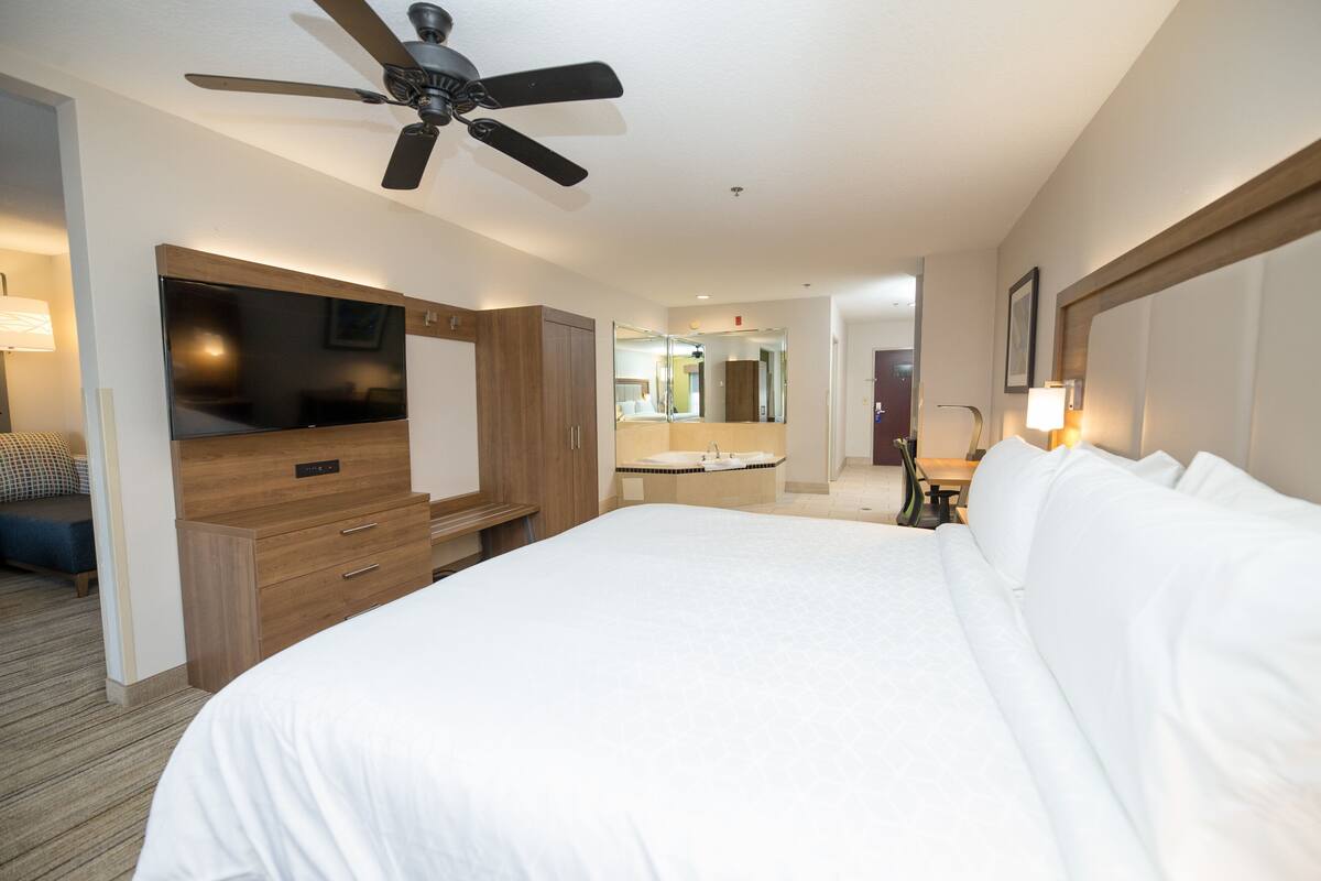 A hotel room with a large bed, a TV, a ceiling fan, a bathroom with a mirror, and a desk with a lamp.