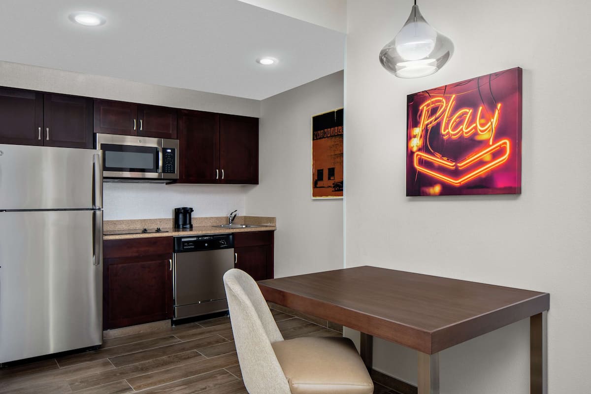 A modern kitchen with stainless steel appliances, dark wooden cabinets, a dining table, and a neon sign on the wall.
