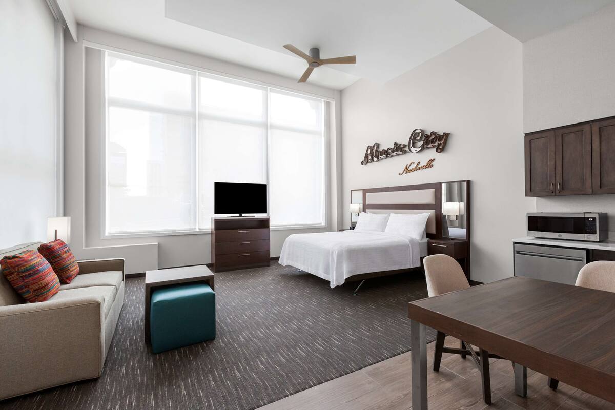 A modern hotel room with a bed, a desk, a television, a kitchenette, and a large window.