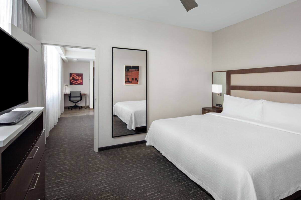A hotel room with a large bed, a desk with a computer, a TV, a mirror, and a door leading to a bathroom and another room.