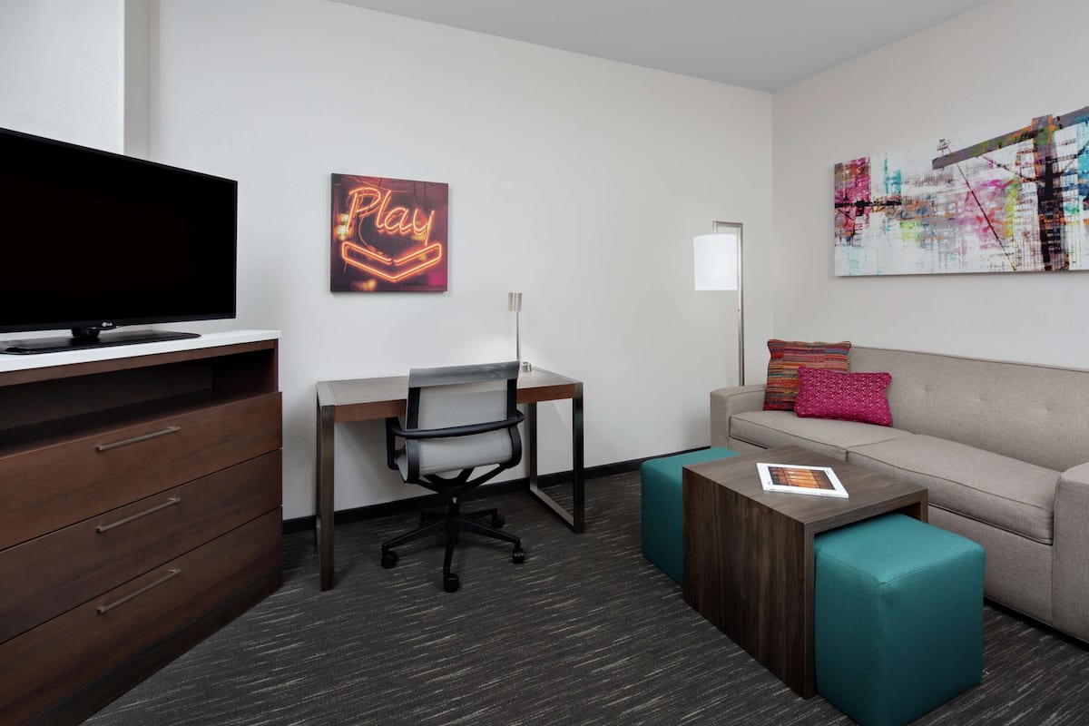 A modern hotel room with a flat-screen TV, a desk with a chair, a sofa with a teal ottoman, and a colorful abstract painting on the wall.