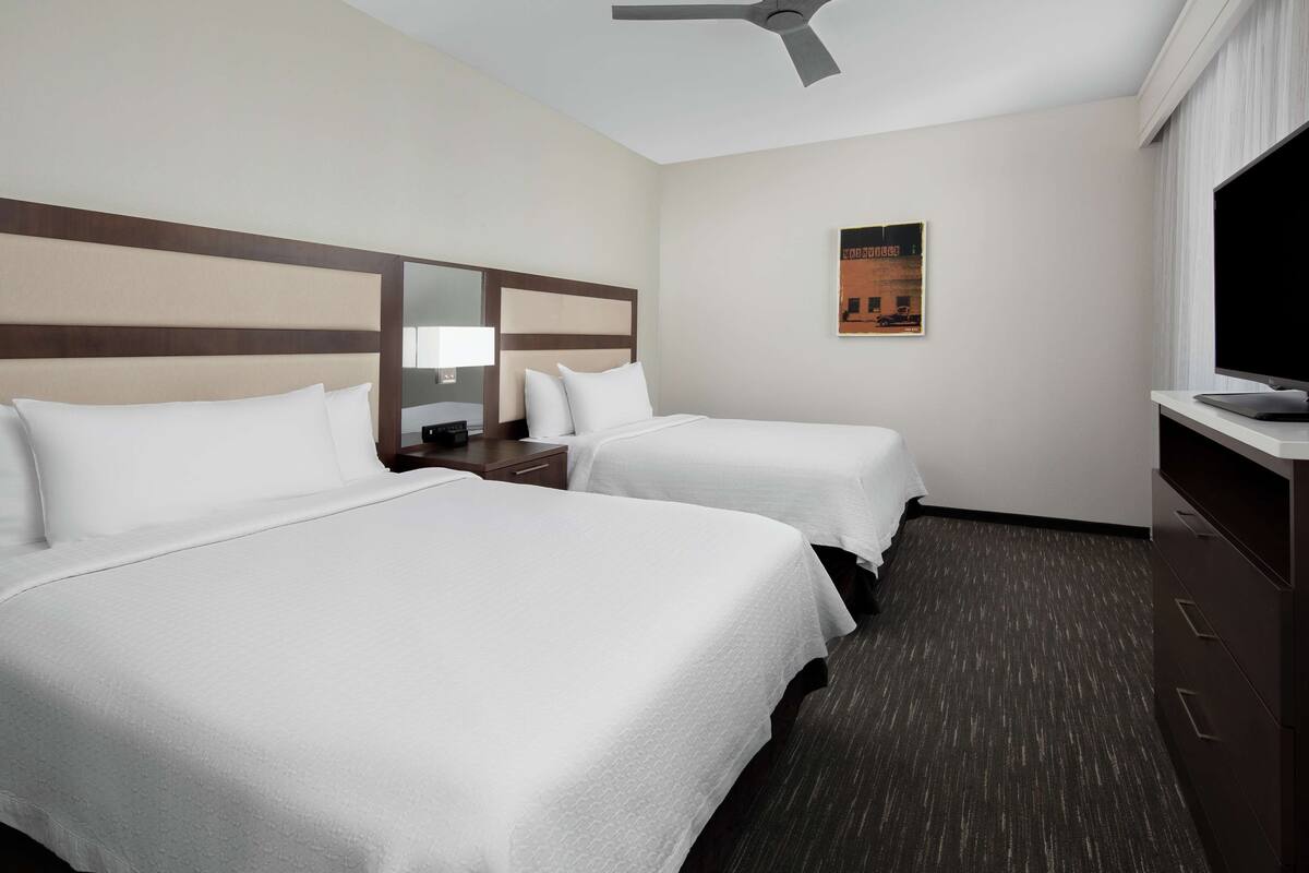 A hotel room with two beds, a ceiling fan, a television, and a poster on the wall.