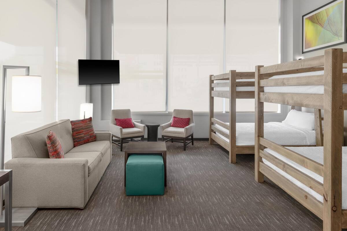 A modern hotel room with a sofa, two armchairs, a small table, a bench, and two bunk beds.