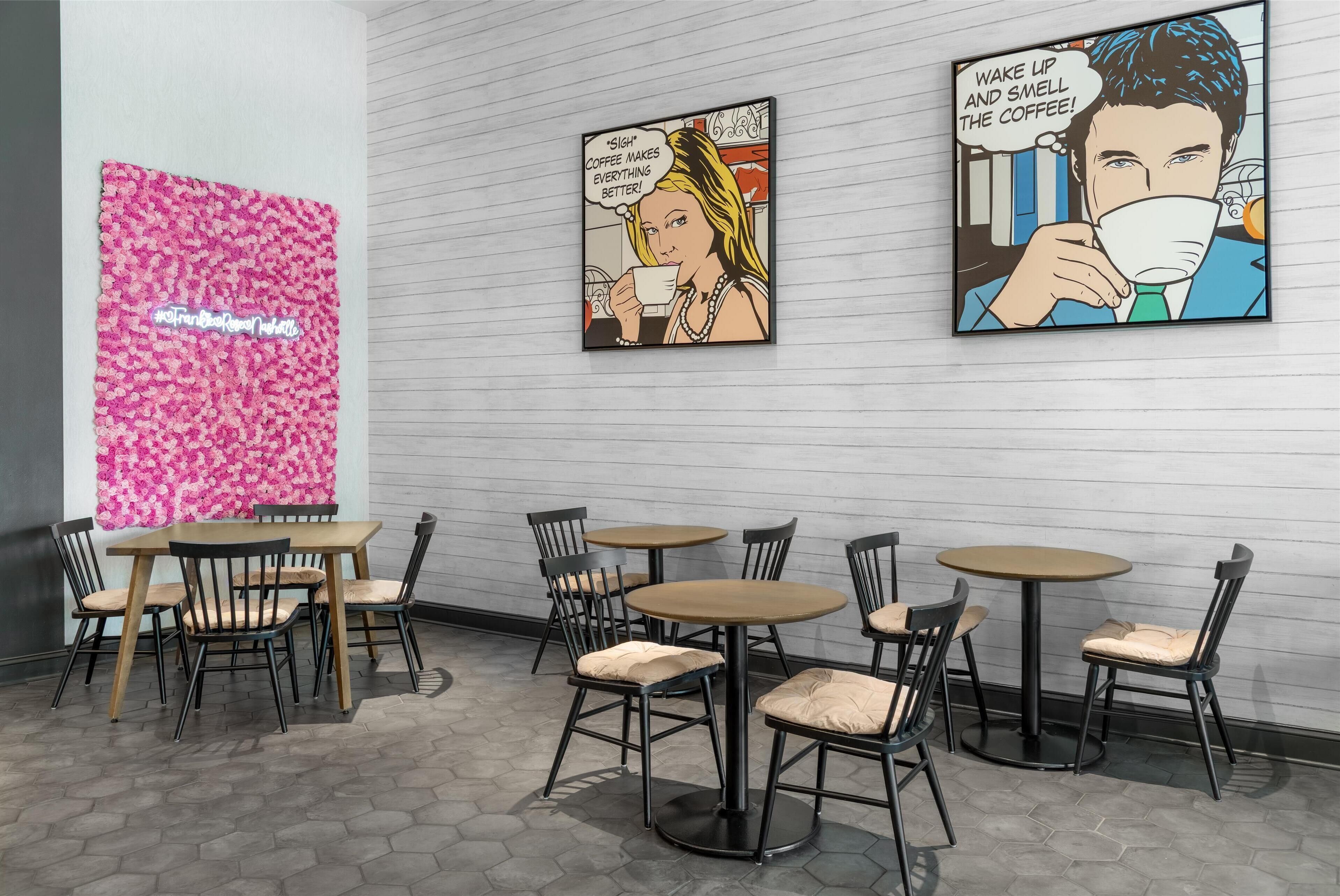 Interior de una cafetería con mesas y sillas, una pared con estampado floral en rosa y dos carteles enmarcados con estilo de cómic.