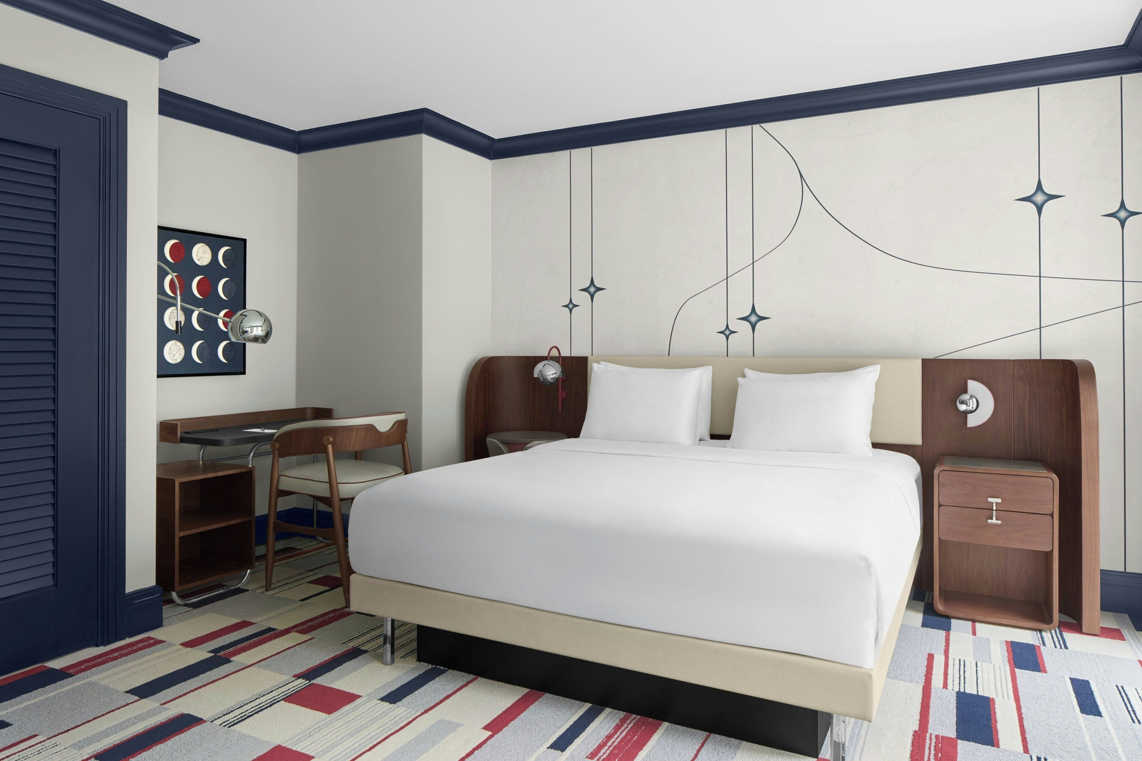A hotel room with a large bed, a desk with a chair, a small bedside table, and a wall-mounted artwork.