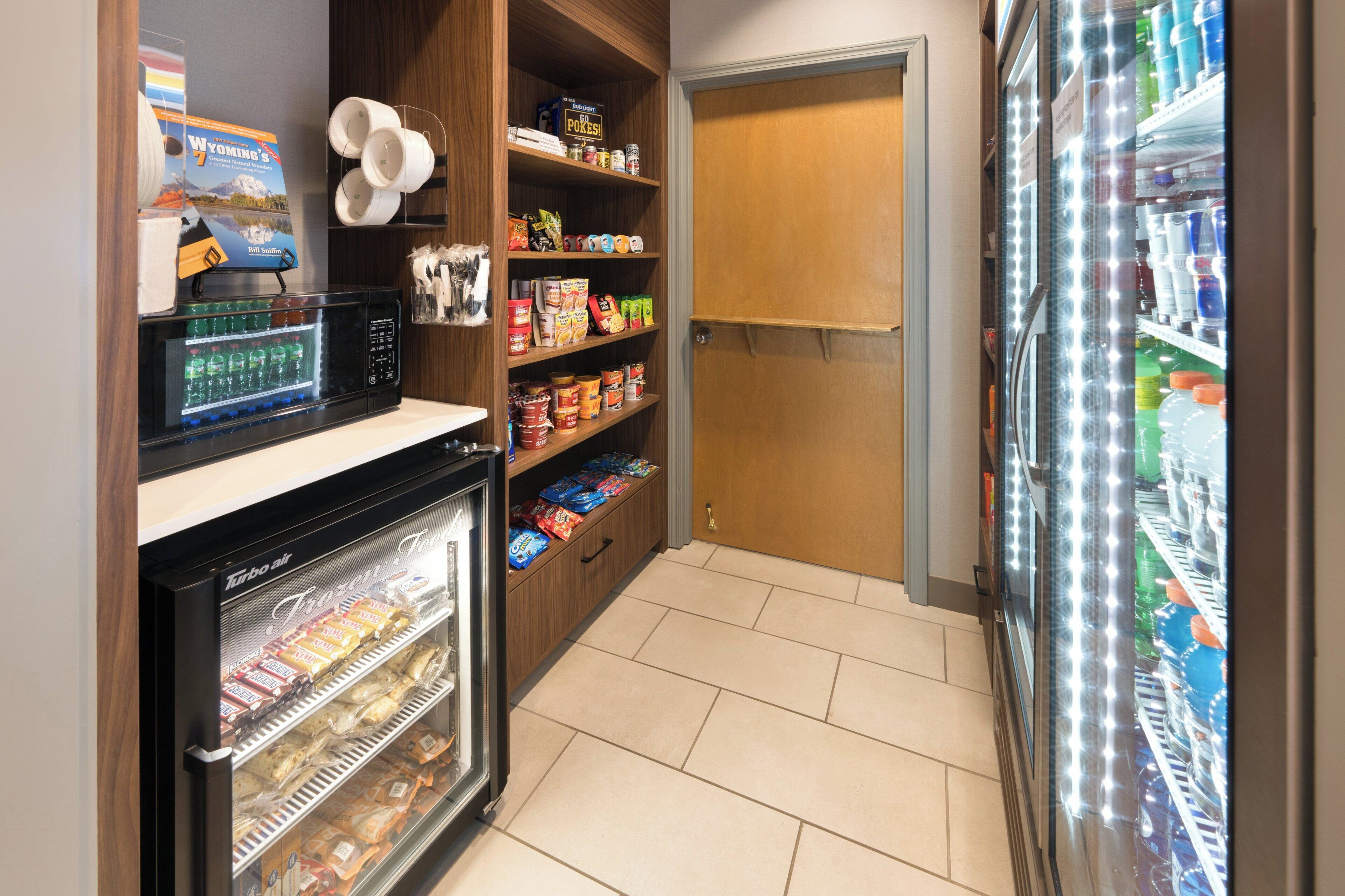 Una kitchenette pequeña con microondas, congelador y refrigerador. Se observan estantes con aperitivos y condimentos.