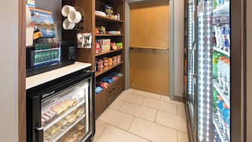 Una kitchenette pequeña con microondas, congelador y refrigerador. Se observan estantes con aperitivos y condimentos.