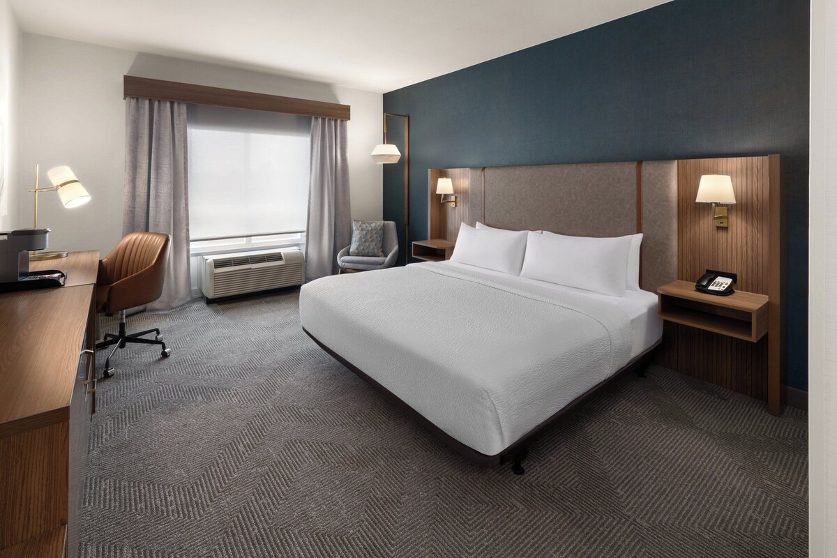 A hotel room with a large bed, a desk, a chair, a desk lamp, a phone, and a window with curtains.