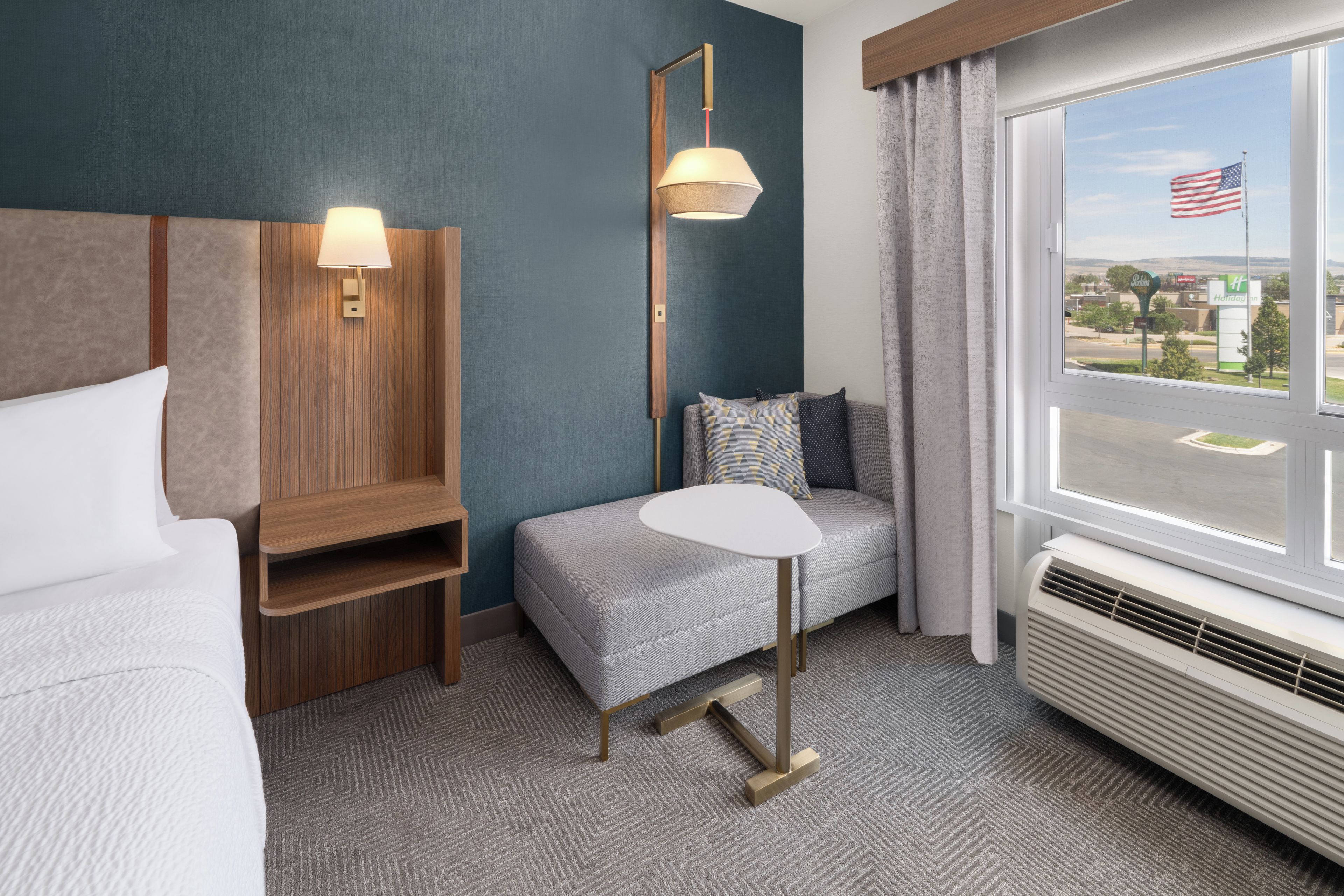 A hotel room with a bed, a small table, a sofa, a lamp, and a window with an American flag.