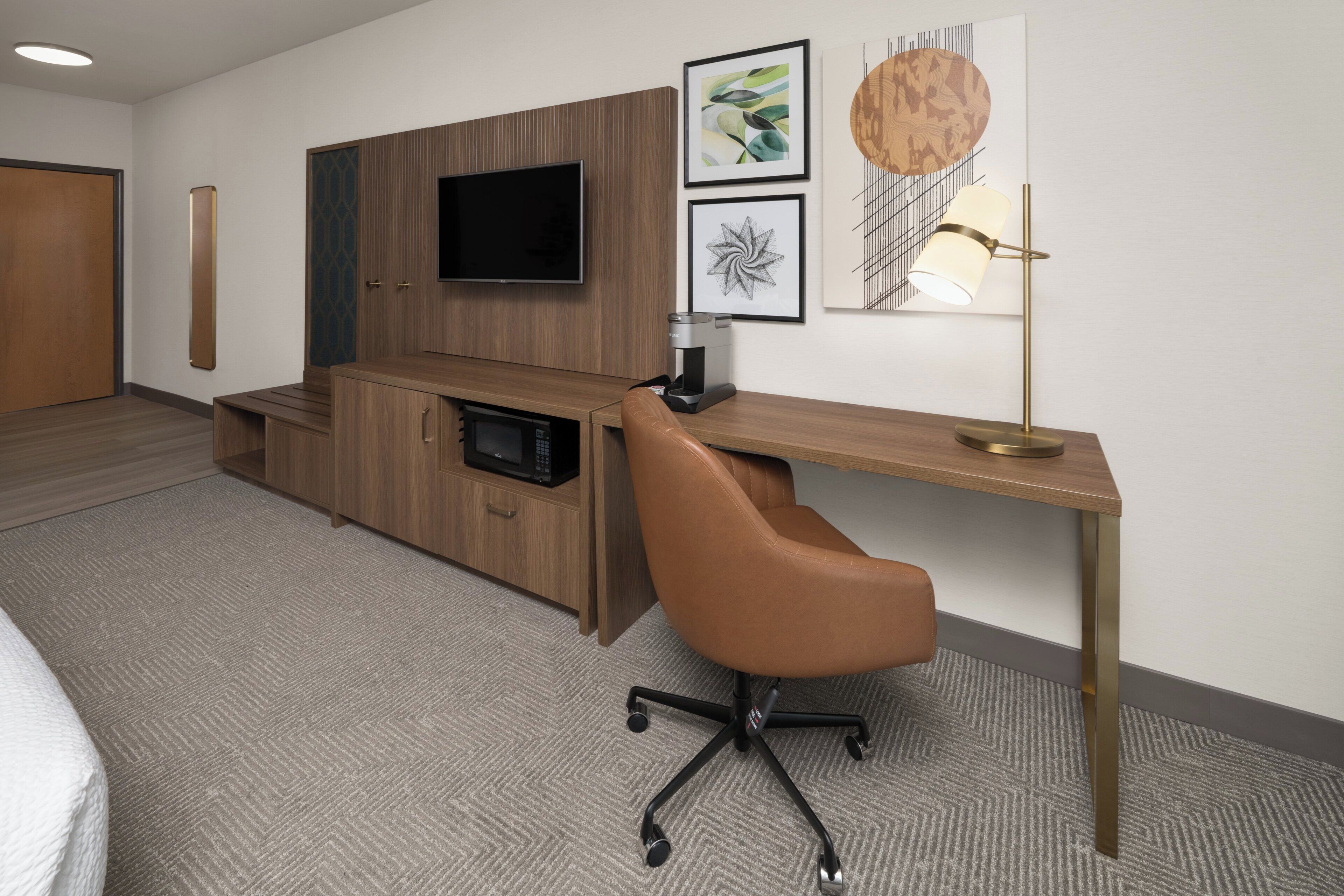 Une chambre d’hôtel avec un bureau en bois, un fauteuil en cuir marron, un téléviseur à écran plat fixé au mur, une machine à café et un tableau d’art abstrait encadré.