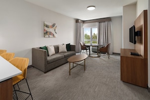 Habitación de hotel con sofá, mesa de comedor, sillas, televisor y ventana con cortinas.