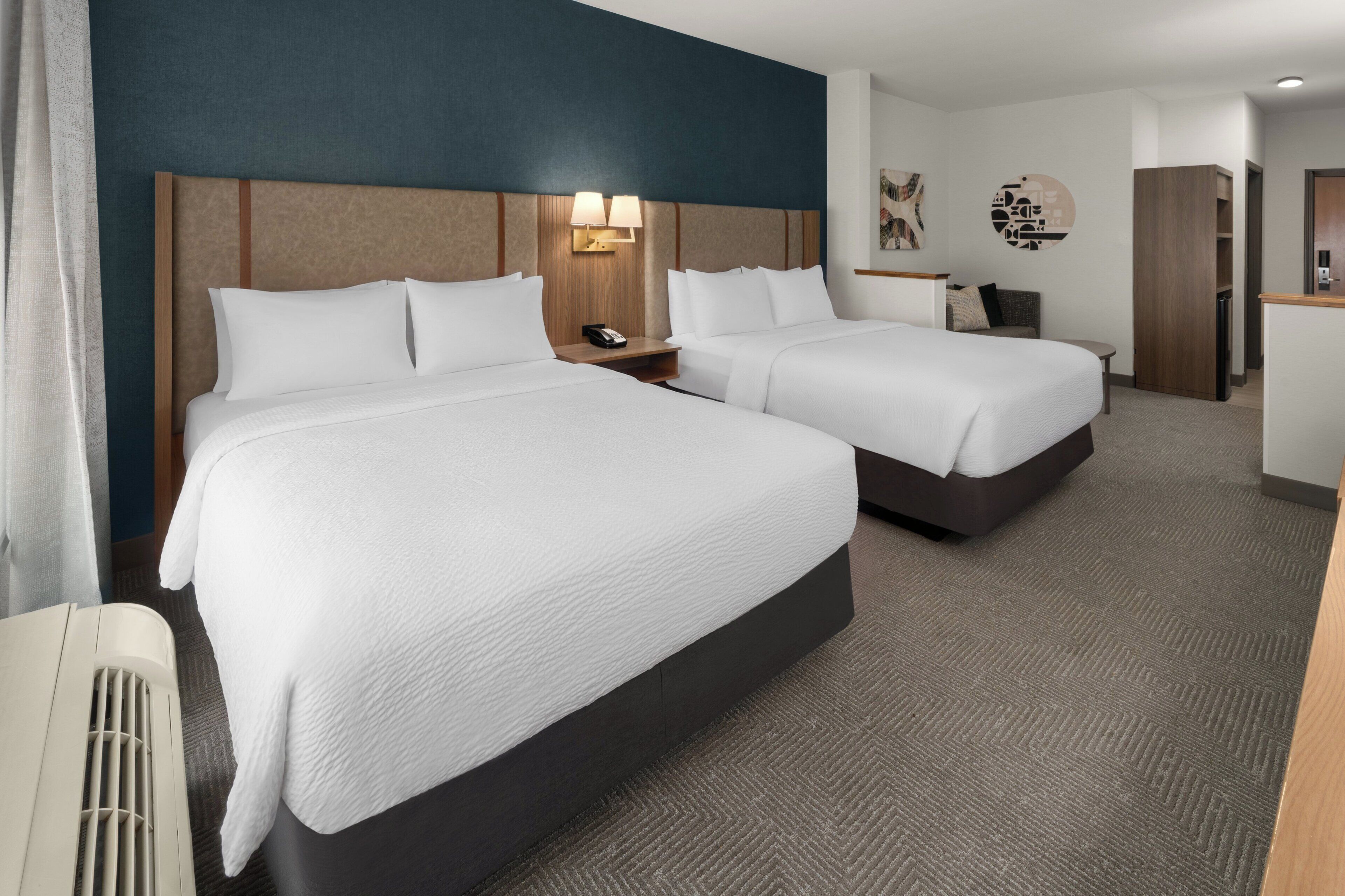 A hotel room with two beds, a bedside table with a lamp, a wall-mounted phone, and a small desk with a chair.