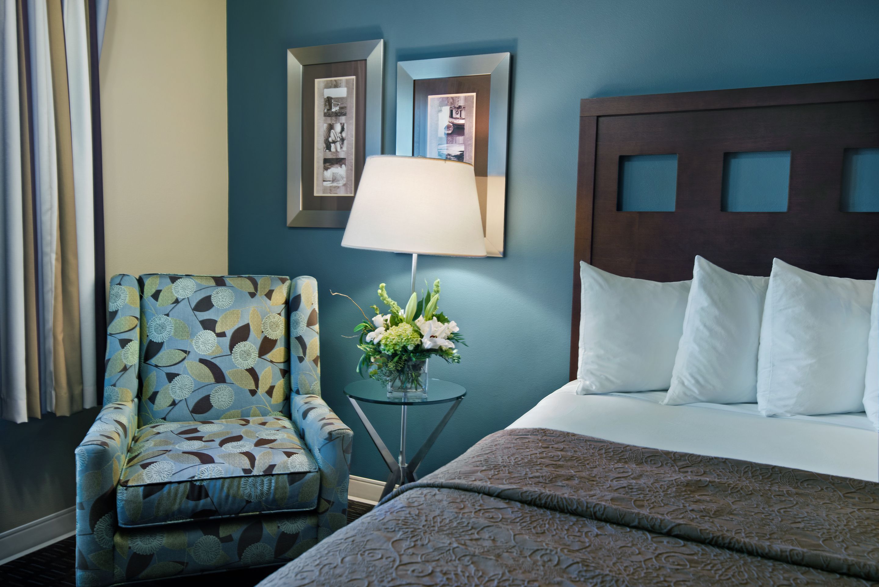 A hotel room with a bed, a chair, a lamp, and a small table with flowers.