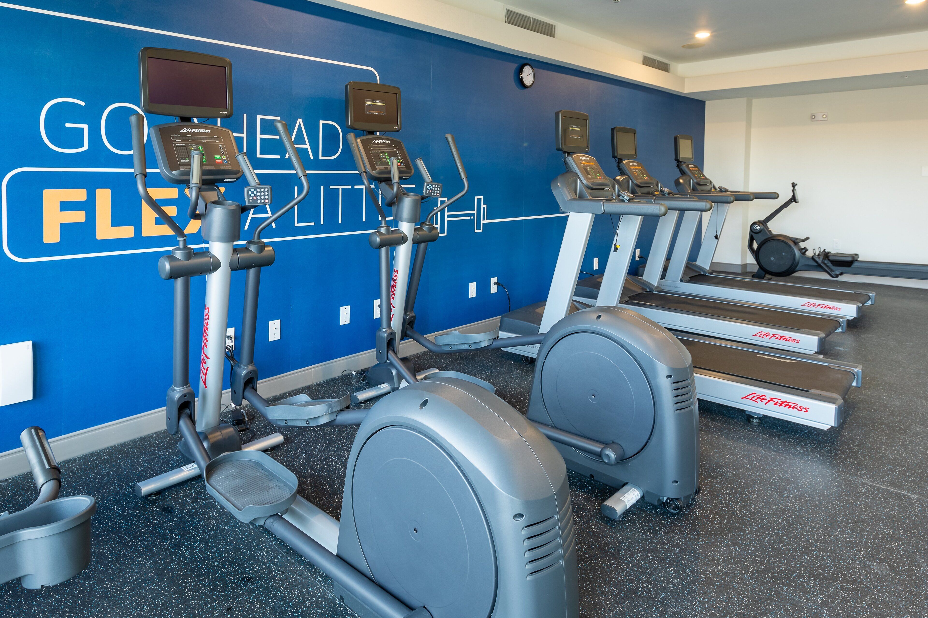 Un gimnasio con varias máquinas de cardio, incluyendo elípticas y cintas de correr, frente a una pared azul con texto motivacional.
