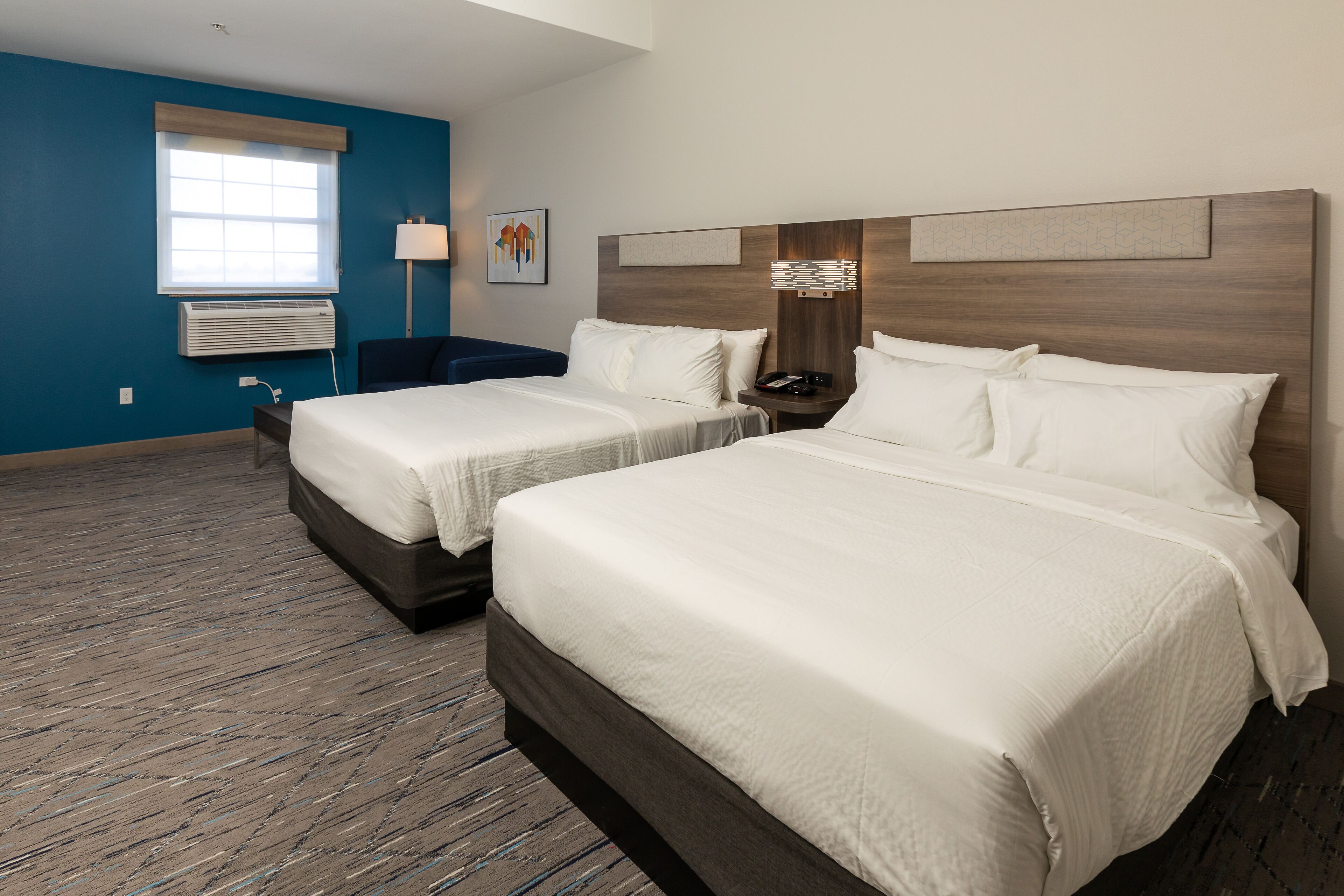 A hotel room with two beds, a window with blinds, a wall-mounted air conditioner, a bedside table with a lamp, and a decorative wall piece.