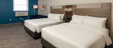 A hotel room with two beds, a window with blinds, a wall-mounted air conditioner, a bedside table with a lamp, and a decorative wall piece.