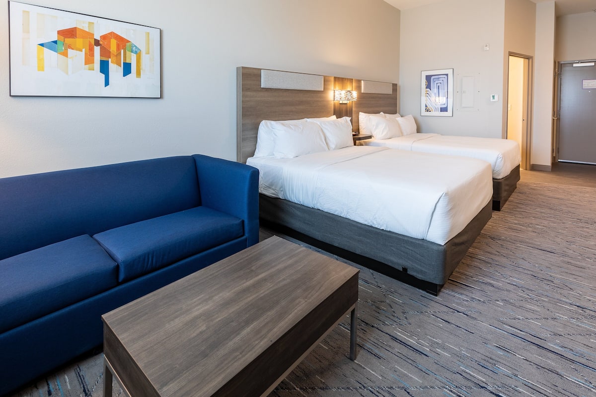 A hotel room with a blue sofa, two beds, a wooden coffee table, and artwork on the walls.