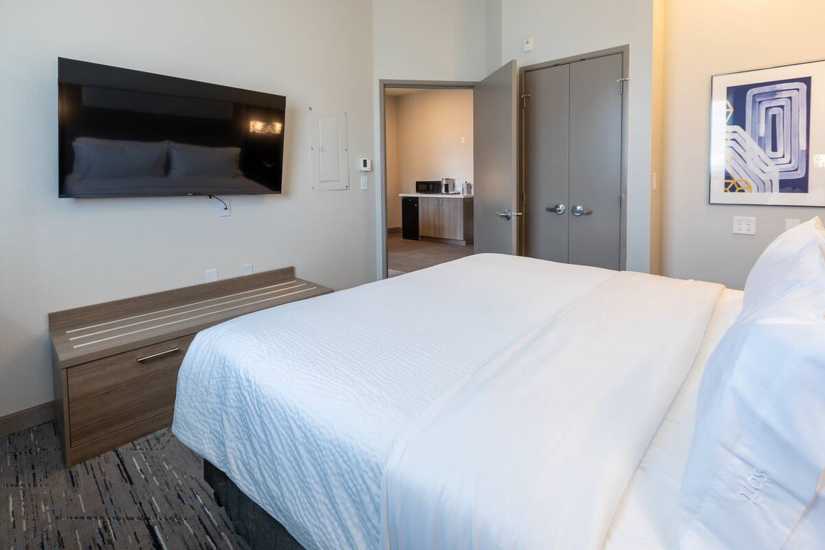 A hotel room with a bed, a TV mounted on the wall, a desk, and a kitchenette in the background.