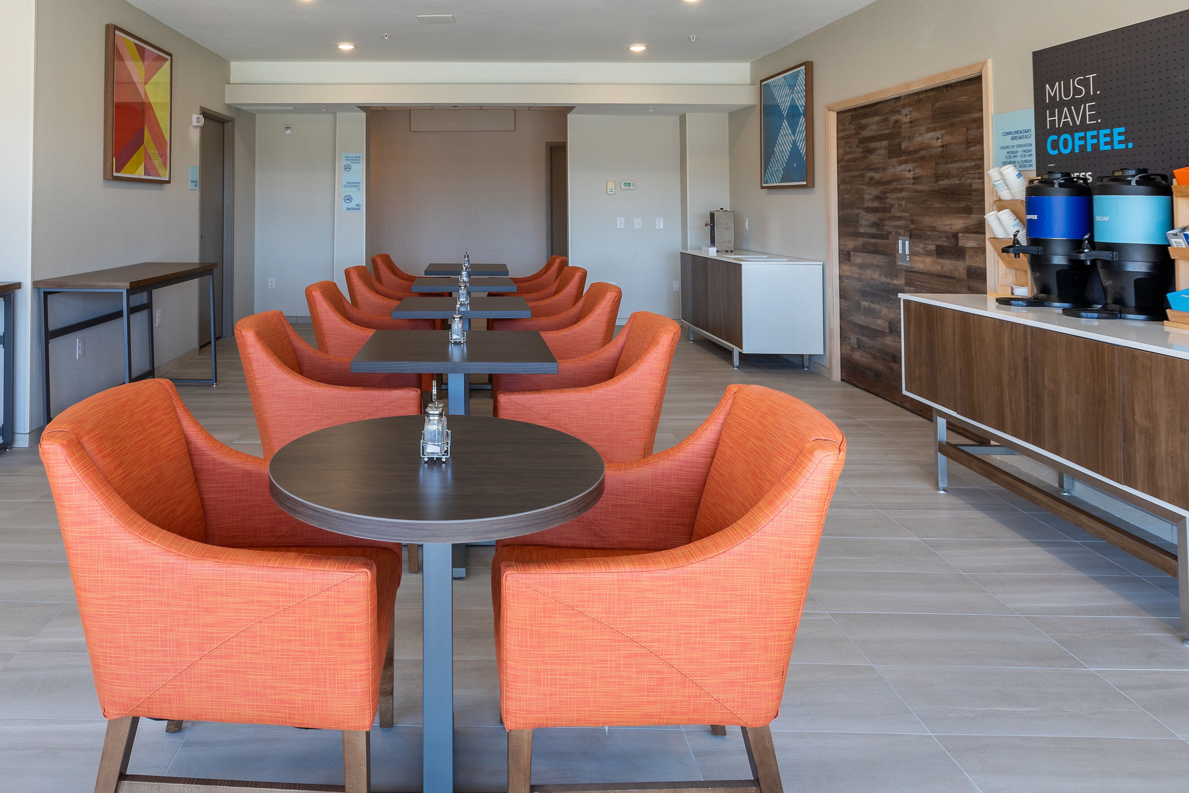 Un espace repas moderne avec des chaises orange et des tables rondes, un coin café équipé de distributeurs et un panneau indiquant : « IL FAUT ABSOLUMENT PRENDRE UN CAFÉ. »
