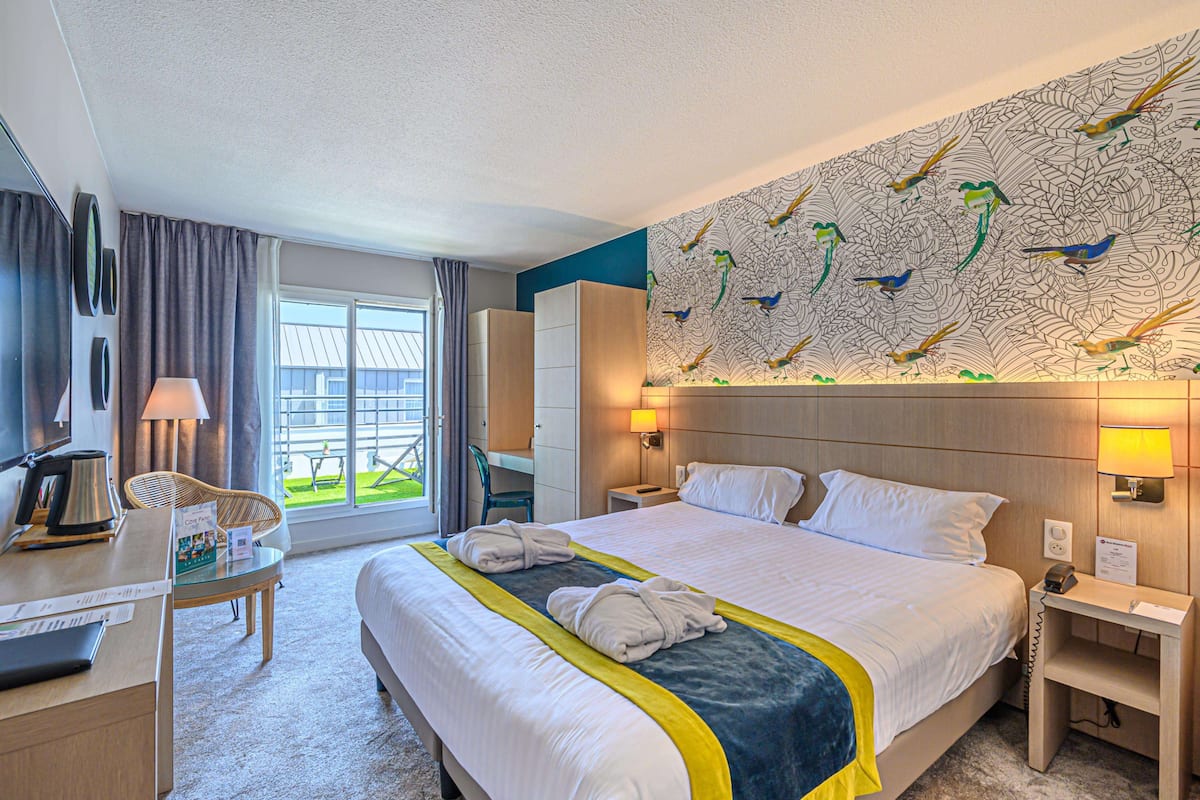 A hotel room with a large bed, a desk with a lamp, a chair, a window with curtains, and a wall with a bird mural.