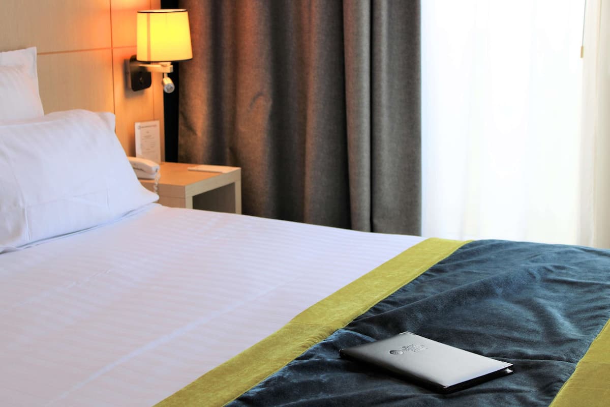 A hotel room with a neatly made bed, a bedside lamp, a telephone, and a laptop on the bed.