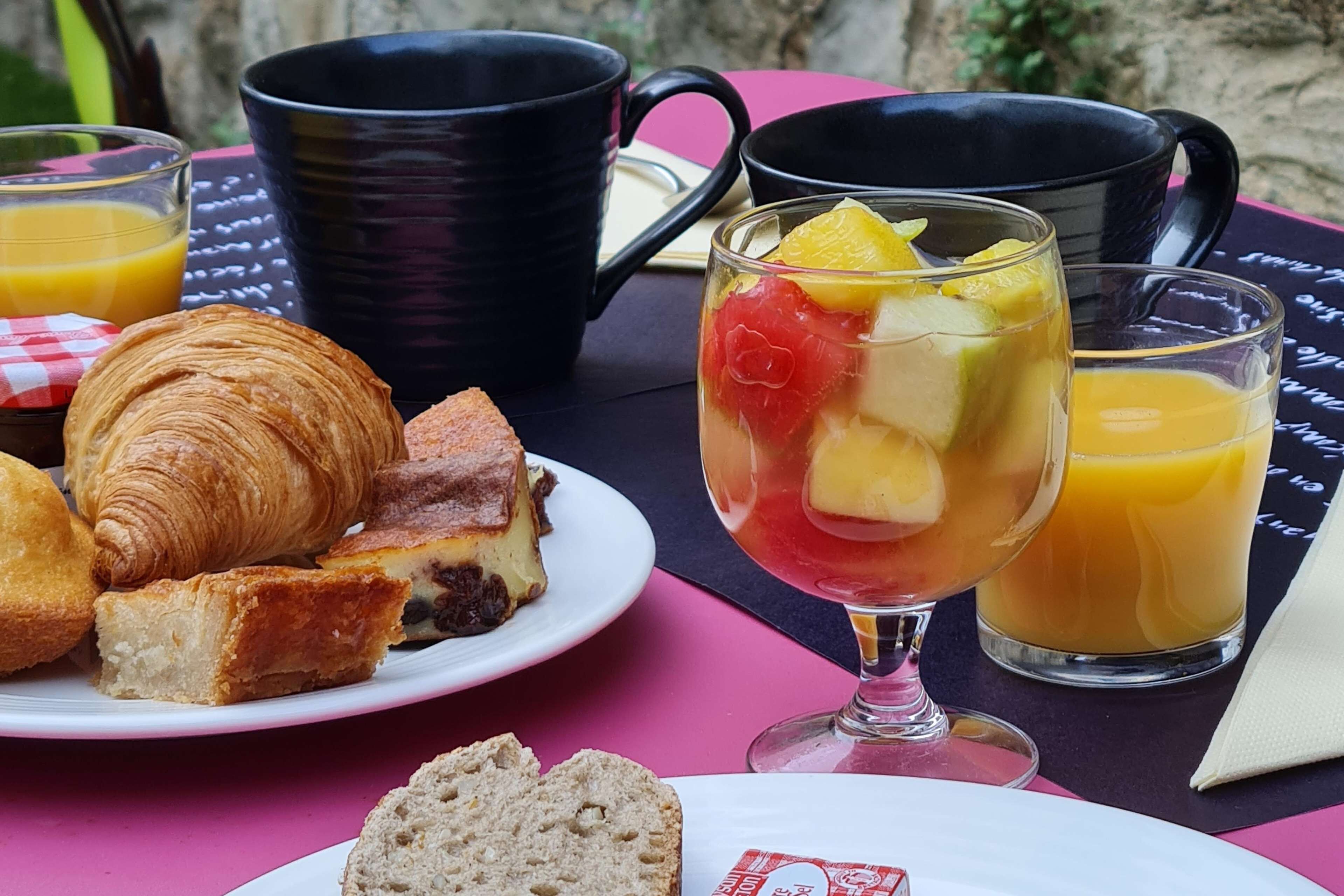 Une table dressée pour le petit-déjeuner, avec des croissants, une salade de fruits et du jus d’orange.